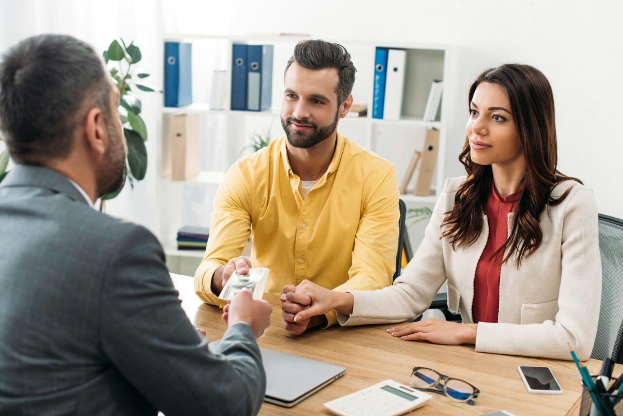 investors-sitting-at-table-and-taking-dollar-banknotes-from-advisor-in-office-e1715620952323